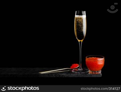 Elegant glass of yellow champagne with red caviar on golden spoon and glass container of caviar on marble board on black background