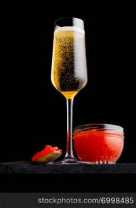 Elegant glass of yellow champagne with red caviar on golden spoon and glass container of caviar on marble board on black.