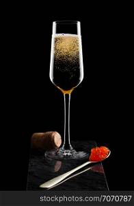 Elegant glass of yellow champagne with red caviar on golden spoon and cork of caviar on marble board on black.