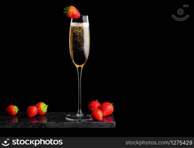 Elegant glass of yellow champagne with strawberry on top and fresh berries on black marble board on black.