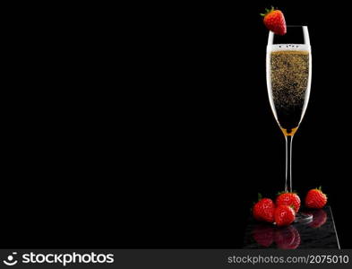 Elegant glass of yellow champagne with strawberry on top and fresh berries on black marble board on black.