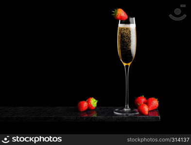 Elegant glass of yellow champagne with strawberry on top and fresh berries on black marble board on black.