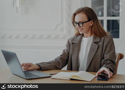 Elegant happy mid adult businesswoman is doing research, browsing internet at home and smiling. Emotional european lady in jacket is freelancer, entrepreneur or accountant. Remote work at the desk.. Elegant businesswoman browsing internet at home and smiling. Remote work at the desk.