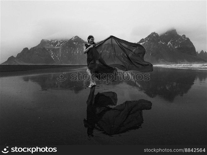 Elegant lady wrapped with chiffon on beach monochrome scenic photography. Picture of person with mountains on background. High quality wallpaper. Photo concept for ads, travel blog, magazine, article. Elegant lady wrapped with chiffon on beach monochrome scenic photography