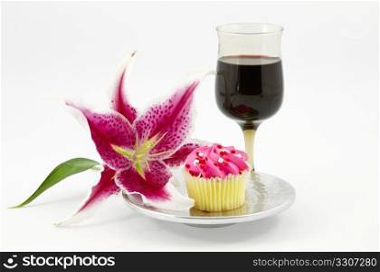 Elegant lily is placed next to cupcake on silver plate accompanied by red wine in stem glass; white background; horizontal;