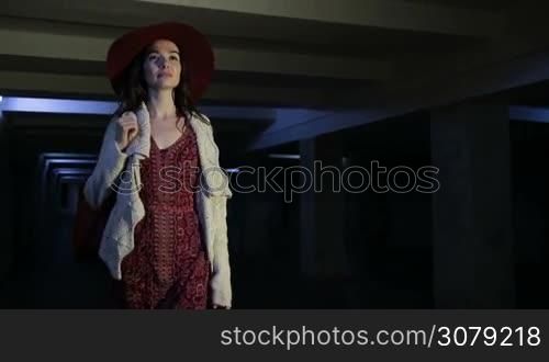 Elegant long brown hair female in fashionable clothes going through dark underground passage alone at night. Sexy young woman walking alone through underground pedestrian crossing in dimmed lights background. Slow motion. Steadicam stabilized shot.