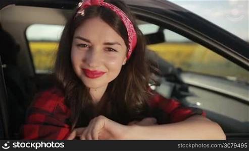 Elegant long brown hair woman in stylish clothes leaning out of car window and smiling. Beautiful pin-up female sitting in the passenger seat and looking out of window while enjoying awesome landscape in countryside. Slo mo. Stabilized shot.