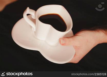 Elegant part body woman in black holding coffee in white heart shaped cup mug on saucer plate. . Woman with cup of coffee