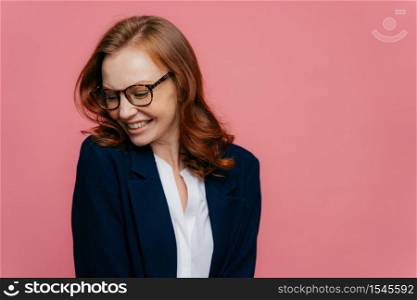 Elegant positive businesswoman focused aside, has sincere smile, wears spectacles, dressed in formal suit, expresses positive emotions, giggles happily, turns from camera, poses in pink studio
