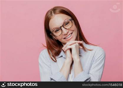 Elegant pretty woman tilts head, keeps hands under chin, wears transparent glasses and shirt, smiles gently, models over pink background. People and positive emotions concept. Charming female