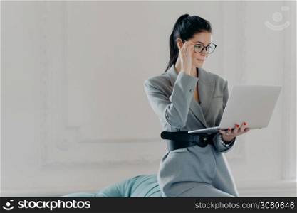 Elegant professional woman banker concentrated at screen of laptop computer, dressed in formal wear, reads documents in online version poses in spacious room, home office. Business, technology concept