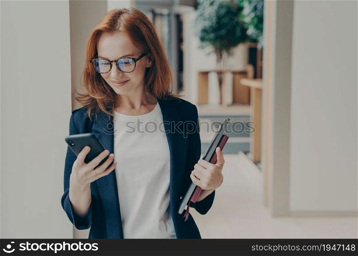 Elegant smiling woman office worker in eyeglasses using modern mobile phone, sending message or chatting with client online, standing in coworking space with laptop and note book in hand. Elegant smiling woman office worker in eyeglasses using modern mobile phone in coworking space