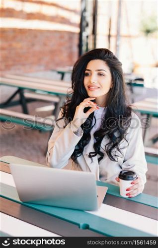 Elegant successful woman freelancer dressed formally using generic laptop computer for remote work, having rest at outdoor cafe drinking delicious coffee having dreamy expression. Lifestyle concept