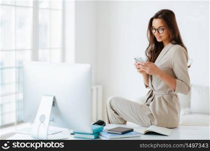 Elegant woman in beige formal suit reads news on website attentively, sits at desktop alone in cabinet, computer monitor and notepads around. Female employee uses modern smartphone in office