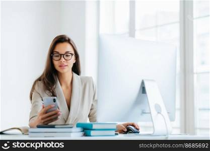 Elegant woman office worker uses mobile phone and computer at one time, sends messages and chats with clients online, monitors news from networks, sits at desktop with pile of notepads and books