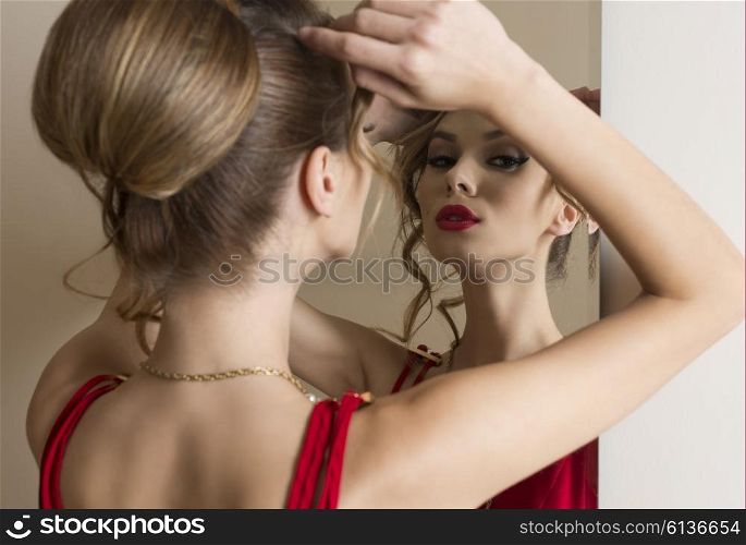 elegant woman with hair-style and make-up looking herself in the mirror and wearing fashion red dress