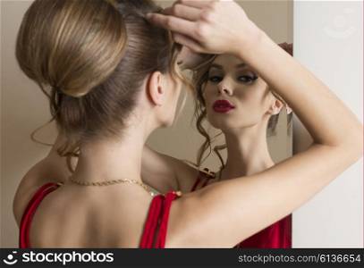 elegant woman with hair-style and make-up looking herself in the mirror and wearing fashion red dress