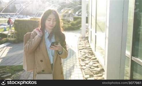 Elegant young asian woman carrying shopping bags over her shoulder and surfing the net with mobile phone in city street after shopping at sunset. Slow motion. Cheerful brunette female using smart phone app to shop online while walking outdoors.