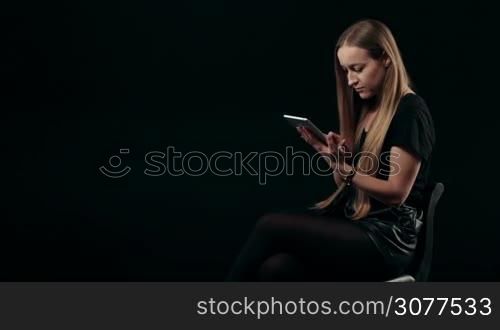 Elegant young blonde female with long hair typing on touchpad and smiling at the camera isolated against black background. Lovely woman working with digital tablet computer and looking at cam with radiant smile.
