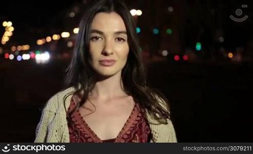 Elegant young brunette with deep brown eyes posing and gazing at camera with mystirous look on night city street. Portrait of sensual woman with long hair playing with cam and looking with shy smile seductively at night. Slow motion. Stabilized shot.