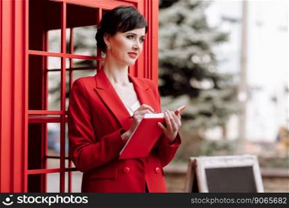 Elegant young businesswoman in red jacket focusing does notes with pen in red notepad outdoors on city street background. Female is feeling happy about both working and having nice rest.. Elegant young businesswoman in red jacket focusing does notes with pen in red notepad outdoors on city street background. Female is feeling happy about both working and having nice rest