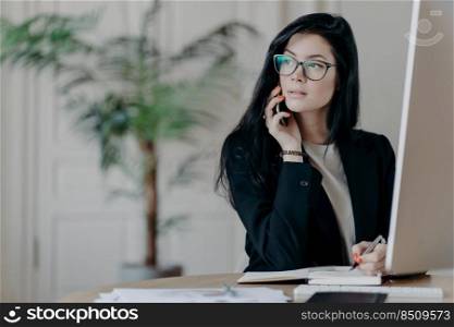 Elegant young woman in formal outfit answers phone call, writes down necessary notes in notepad, looks away with thoughtful expression, works at modern work place, has telephone conversation
