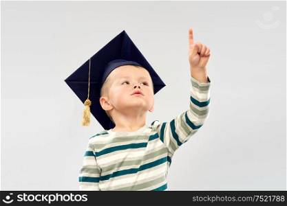 elementary school, preschool education and childhood concept - portrait of little boy in mortar board pointing finger up over grey background. little boy in mortar board pointing finger up