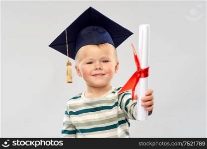 elementary school, preschool education and childhood concept - portrait of smiling little boy in mortar board with diploma over grey background. smiling little boy in mortar board with diploma