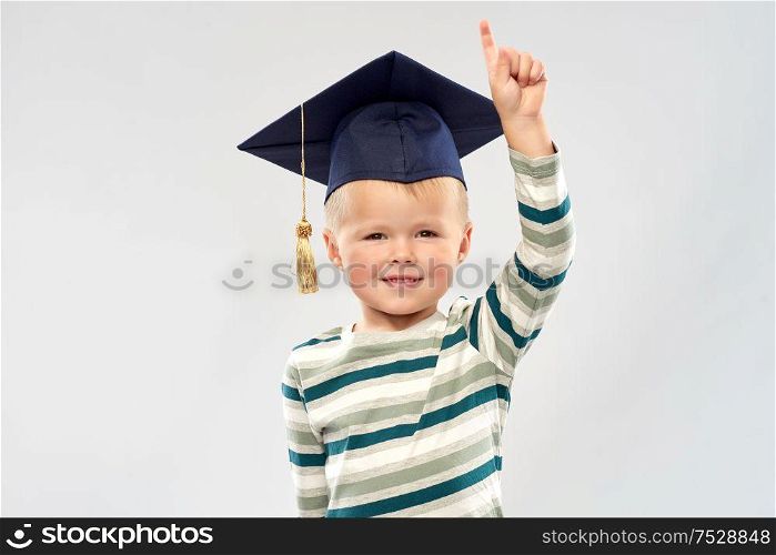 elementary school, preschool education and childhood concept - portrait of smiling little boy in mortar board over grey background. little boy in mortar board pointing finger up