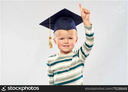 elementary school, preschool education and childhood concept - portrait of smiling little boy in mortar board over grey background. little boy in mortar board pointing finger up