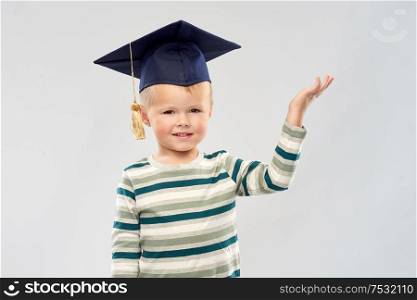 elementary school, preschool education and childhood concept - portrait of smiling little boy in mortar board holding something in empty hand over grey background. little boy in mortar board with empty hand