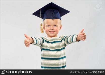 elementary school, preschool education and childhood concept - portrait of smiling little boy in mortar board showing thumbs up over grey background. happy little boy in mortar board showing thumbs up