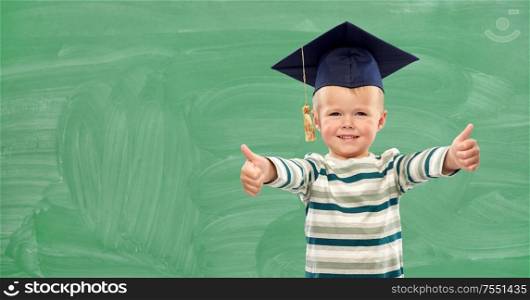 elementary school, preschool education and childhood concept - portrait of smiling little boy in mortar board showing thumbs up over green chalk board background. happy little boy in mortar board showing thumbs up