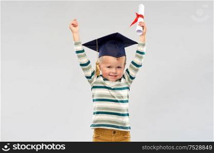 elementary school, preschool education and success concept - portrait of smiling little boy in mortar board with diploma celebrating triumph over grey background. smiling little boy in mortar board with diploma