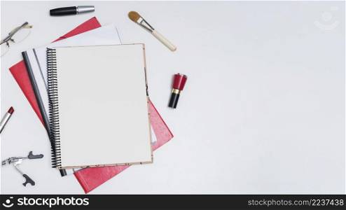 elevated view blank spiral notebook surrounded by nail varnish makeup brush mascara eyeglass lipstick eyelash curler white background