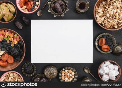 elevated view blank white paper surrounded with delicious dried fruits nuts sweets ramadan black textured backdrop