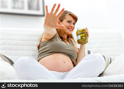 Embarrassed beautiful pregnant woman sitting on sofa at home and holding jar of pickles in hands&#xA;