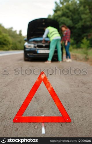 Emergency stop sign, young couple at the opened hood on road, car breakdown. Broken automobile or emergency accident with vehicle, trouble with engine on highway