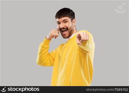 emotion and people concept - happy smiling young man in yellow sweatshirt making phone call gesture and pointing finger to camera over grey background. man making phone call gesture and pointing finger