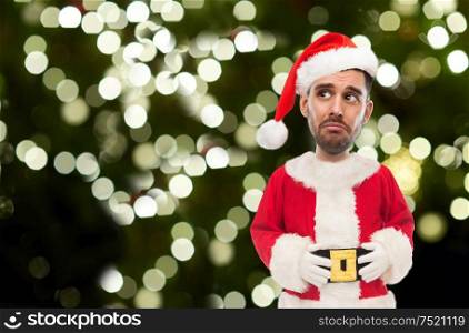 emotion, christmas and winter holiday concept - sad man in santa claus costume over festive lights on dark green background (funny cartoon style character with big head). sad man in santa claus costume over christmas tree