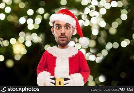 emotion, christmas and winter holiday concept - surprised man in santa claus costume over festive lights on dark green background (funny cartoon style character with big head). surprised man in santa costume over christmas tree