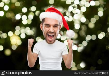 emotion, christmas and winter holiday concept - young man in santa hat celebrating victory and screaming over festive lights on dark green background (funny cartoon style character with big head). man in santa hat celebrating victory at home