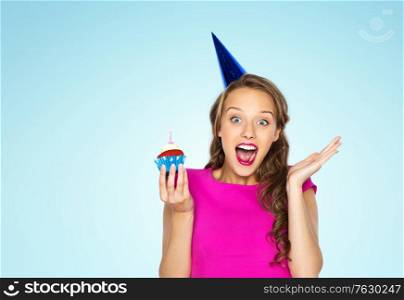 emotion, expression and celebration concept - happy young woman or teen girl in pink dress and party cap with birthday cupcake over blue background. happy woman or teen girl with birthday cupcake