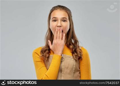 emotion, expression and people concept - scared or yawning teenage girl closing her mouth by hand over grey background. teenage girl closing her mouth by hand or yawning