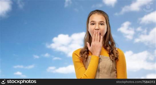 emotion, expression and people concept - scared or yawning teenage girl closing her mouth by hand over blue sky and clouds background. teenage girl closing her mouth by hand or yawning
