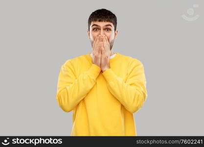 emotion, expression and people concept - shocked and speechless young man in yellow sweatshirt covering his mouth with hands over grey background. shocked young man covering his mouth with hands