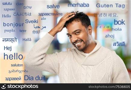 emotion, expression and people concept - smiling indian man in knitted woolen sweater touching his hair over greeting words in different foreign languages. indian man in knitted sweater touching his hair