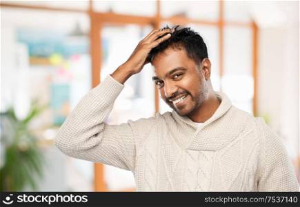 emotion, expression and people concept - smiling indian man in knitted woolen sweater touching his hair over office background. indian man in knitted sweater touching his hair
