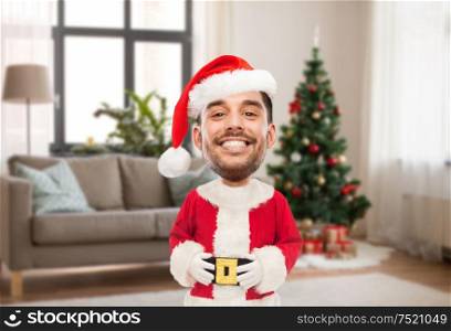 emotion, expression and winter holiday concept - happy smiling young man in santa claus costume over christmas tree on home background (funny cartoon style character with big head). smiling man in santa costume over christmas tree