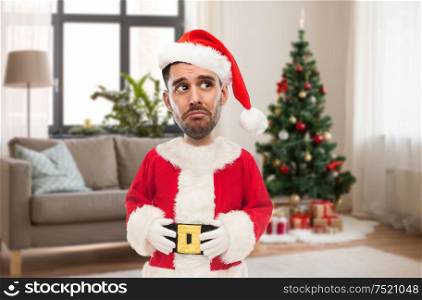 emotion, expression and winter holiday concept - sad man in santa claus costume over christmas tree on home background (funny cartoon style character with big head). sad man in santa claus costume over christmas tree